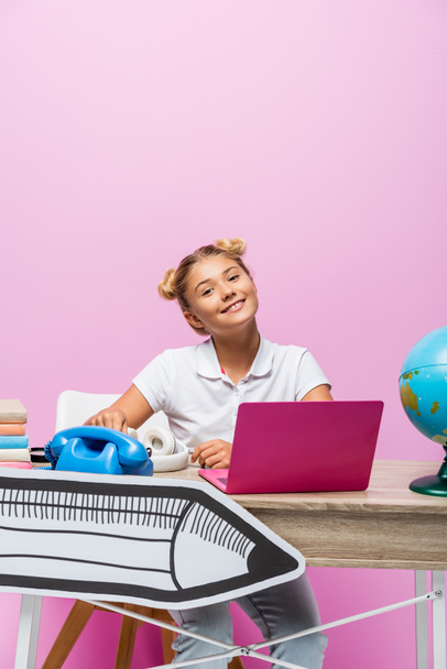 Child sitting near laptop, globe and paper art on desk on pink background - Foto, Imagen