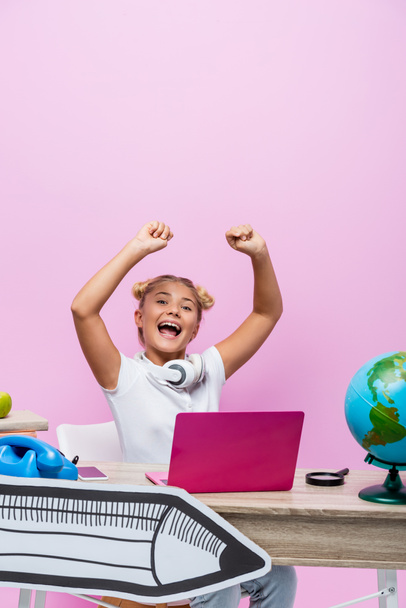 Excited schoolkid sitting near laptop, globe and paper art on pink background - Fotó, kép