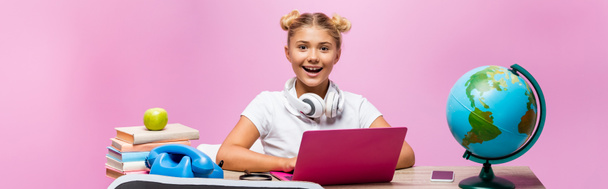 Horizontal crop of child in headphones sitting near gadgets, globe and books on pink background - Photo, Image