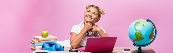 Panoramic shot of schoolgirl in headphones sitting near gadgets, books and globe on pink background - 写真・画像