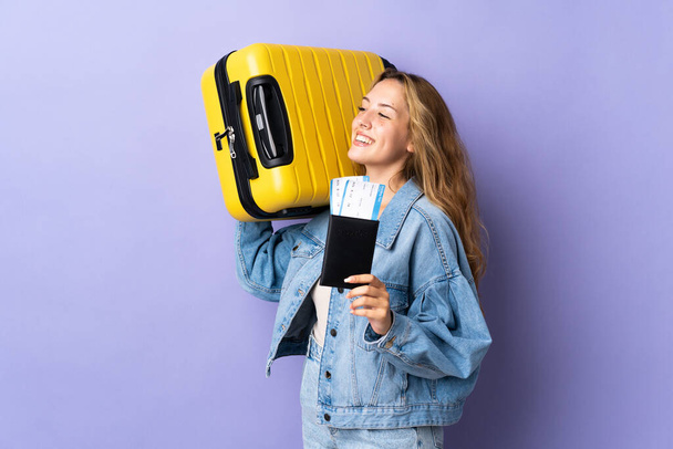 Young blonde woman isolated on purple background in vacation with suitcase and passport - 写真・画像