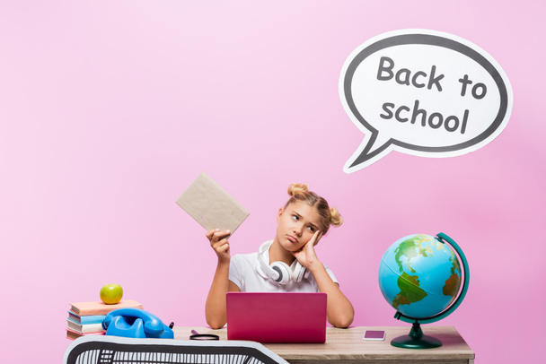 Tired kid holding book near gadgets, globe and speech bubble with back to school lettering on pink background - Foto, Imagen