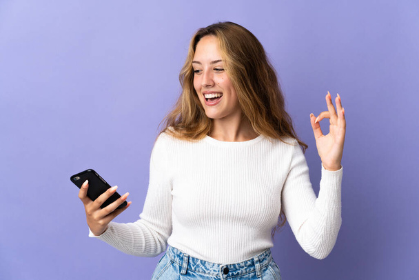Jeune femme blonde isolée sur fond violet en utilisant le téléphone mobile et faire OK signe - Photo, image