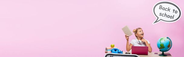 Panoramic shot of kid holding book near globe, gadgets and paper artwork on pink background - Фото, зображення