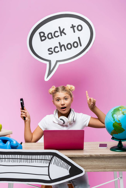 Schoolgirl with magnifying glass pointing at paper art near gadgets, globe and books on pink background - Fotoğraf, Görsel