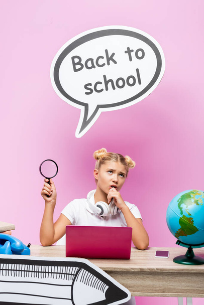 Pensive schoolgirl with magnifying glass near gadgets, books and paper art on pink background - Photo, Image