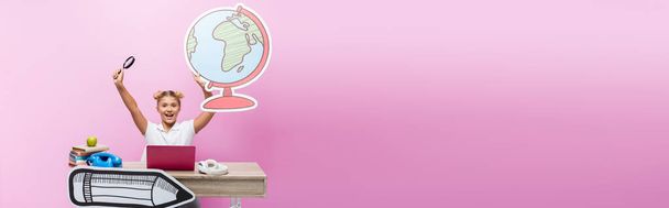 Panoramic shot of excited schoolgirl holding paper globe and magnifying glass near laptop and books on pink background - 写真・画像
