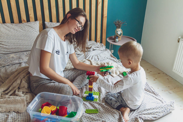 Mom and kid play together in the bedroom on a bed with plastic blocks - Zdjęcie, obraz