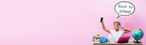 Foto panorámica de colegiala sosteniendo teléfono inteligente y lupa cerca del globo y burbuja del habla con letras de vuelta a la escuela sobre fondo rosa - Foto, Imagen