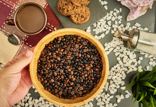 Mix of natural and roasted coffee in a bowl held by a caucasian hand on wooden table with cookies, cup of coffee, spoon and sugar - Photo, Image
