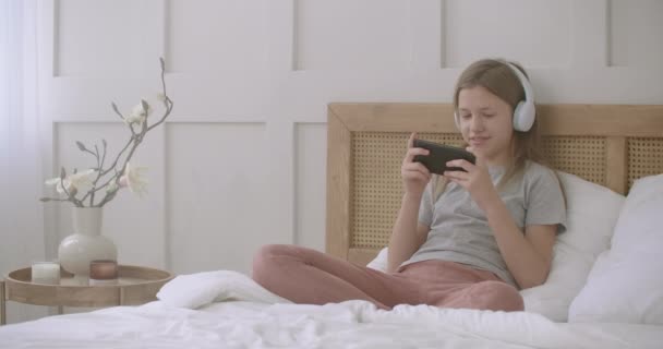 dziecko dziewczyna jest gra w smartfona po lekcjach szkolnych, siedzi w łóżku sam w domu, dzieciństwo - Materiał filmowy, wideo