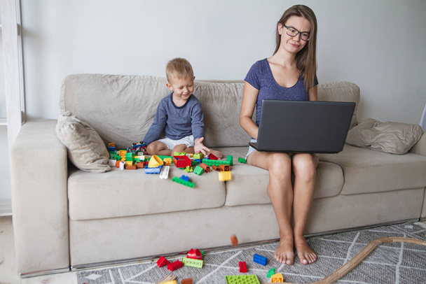 Little kid plays on sofa while mom works with laptop - Foto, imagen