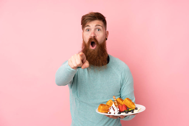 Redhead man with long beard holding waffles over isolated pink background surprised and pointing front - Fotografie, Obrázek