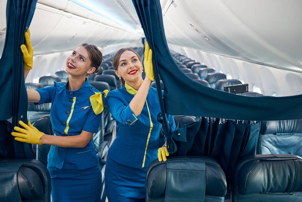 Beautiful females air hostesses in blue uniform with yellow scarf and gloves preparing plane to the passenger - Photo, Image