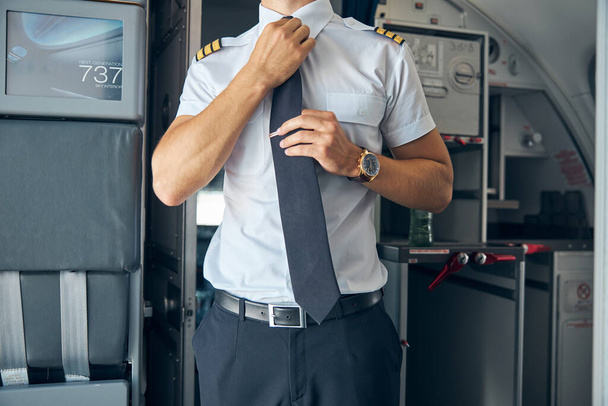 Genç erkek kaptan kravatını uçakta düzeltiyor. - Fotoğraf, Görsel