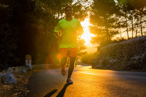 Primer plano del joven corriendo por un camino con grandes pinos alrededor con hermosa puesta de sol dorada en el fondo, el hombre está usando ropa deportiva y llamativo - Foto, imagen