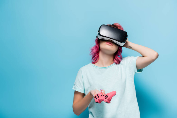 KYIV, UKRAINE - JULY 29, 2020: young woman with pink hair in vr headset with joystick on blue background - Фото, изображение