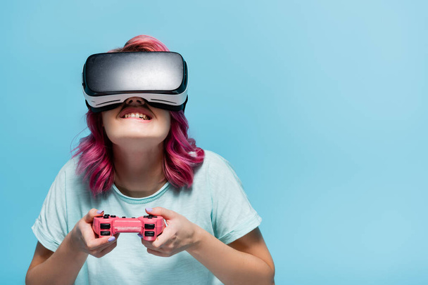 KYIV, UKRAINE - JULY 29, 2020: concentrated young woman with pink hair in vr headset playing video game with joystick isolated on blue - Fotó, kép