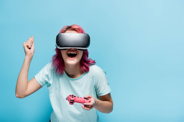 KYIV, UKRAINE - JULY 29, 2020: excited young woman with pink hair in vr headset with joystick on blue background - Fotografie, Obrázek