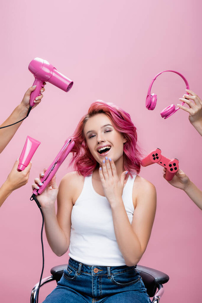 KYIV, UKRAINE - JULY 29, 2020: young woman with colorful hair near straightener, headphones, cosmetic cream, joystick and hairdryer in hands isolated on pink - Foto, Imagem