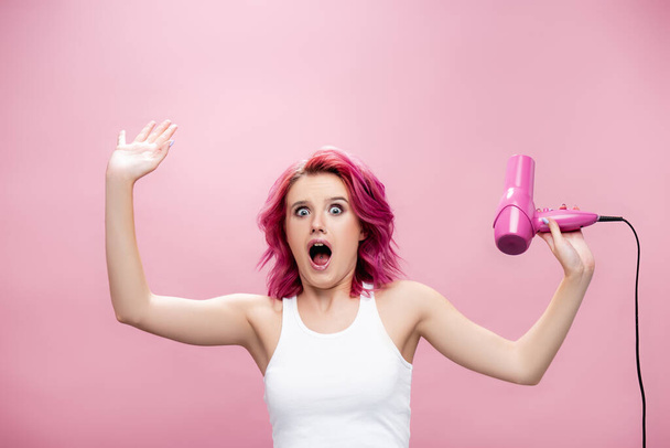 verängstigte junge Frau mit buntem Haar und Haartrockner isoliert auf rosa - Foto, Bild