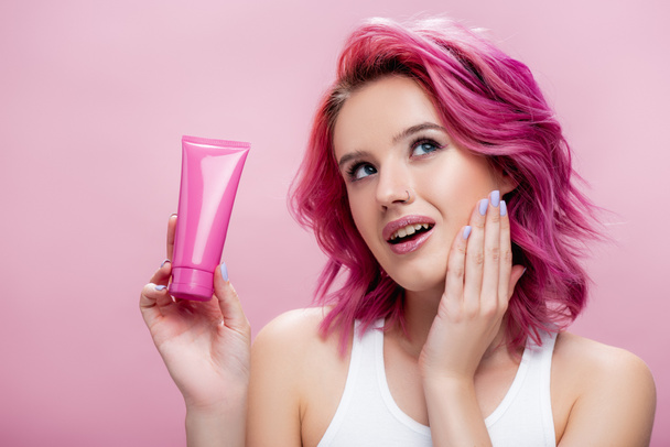 verträumte junge Frau mit bunten Haaren, die eine Tube Kosmetikcreme auf rosa - Foto, Bild