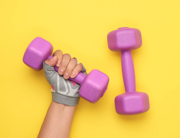 kobieta ręka w różowej rękawicy sportowej posiada fioletowy jeden kilogram hantle na żółtym tle - Zdjęcie, obraz