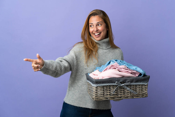 Frau mittleren Alters hält einen Wäschekorb isoliert auf lila Hintergrund zeigt mit dem Finger zur Seite und präsentiert ein Produkt - Foto, Bild