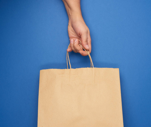 main féminine tient un sac en papier brun vide par les poignées sur un fond bleu, rejet de sacs en plastique, zéro déchet  - Photo, image