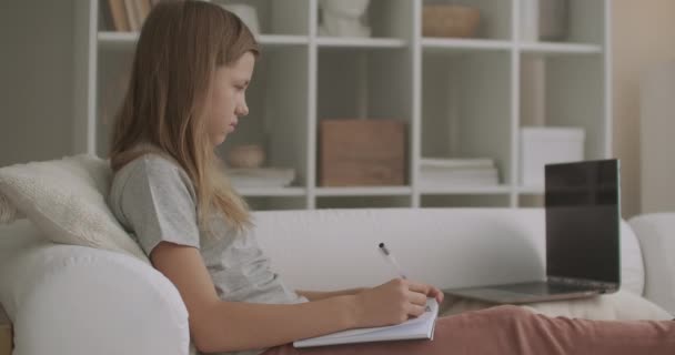 nastolatka robi pracę domową w pokoju mieszkania po szkole, pisze w zeszycie ćwiczeń, siedzi na kanapie, e-learning dla dzieci w wieku szkolnym - Materiał filmowy, wideo