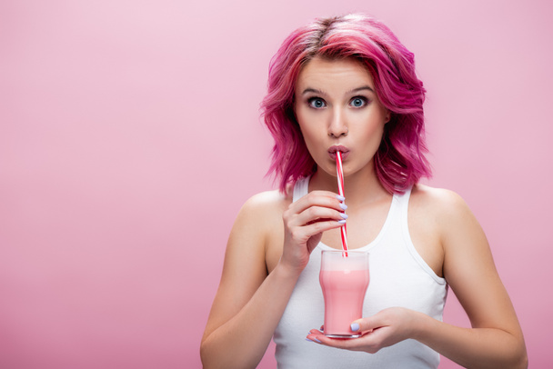 überraschte junge Frau mit buntem Haar trinkt Erdbeer-Milchshake aus Stroh isoliert auf rosa - Foto, Bild