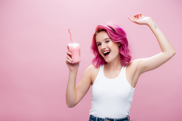mujer joven con pelo colorido sosteniendo batido de fresa en vidrio con paja para beber y bailando aislado en rosa - Foto, Imagen