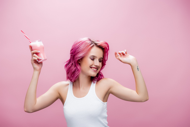 young woman with colorful hair holding strawberry milkshake in glass with drinking straw and dancing isolated on pink - Fotografie, Obrázek