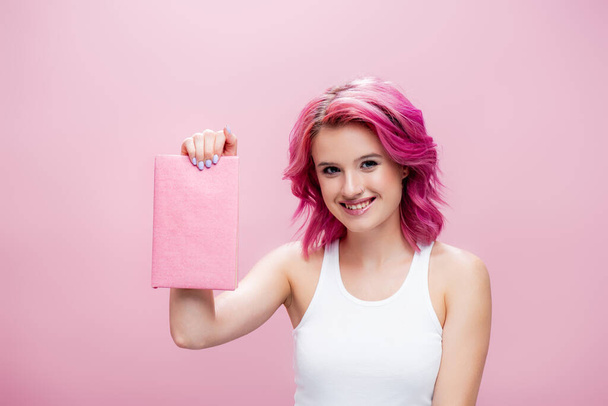 молодая женщина с красочными волосами представляет книгу, изолированную на розовый - Фото, изображение