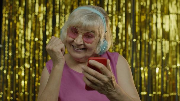 Elegante anciana feliz sosteniendo el teléfono celular, escuchando música en los auriculares y bailando - Foto, imagen
