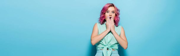 шокированная молодая женщина с розовыми волосами, покрывающими рот на синем фоне, панорамный снимок - Фото, изображение