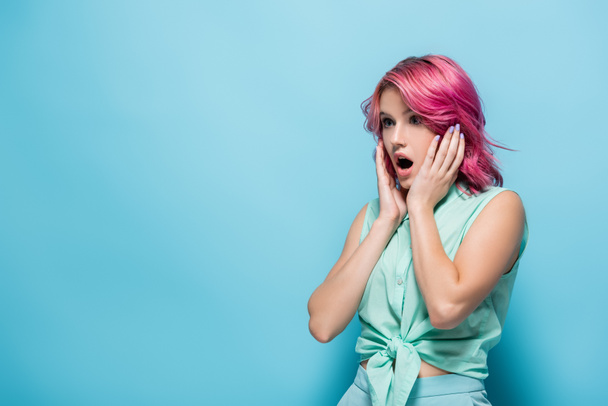mujer joven sorprendida con el pelo rosa y la boca abierta tocando la cara sobre fondo azul - Foto, Imagen