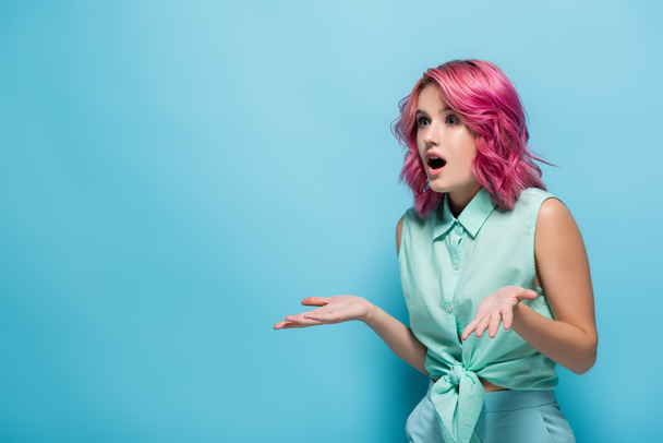 mujer joven sorprendida con el pelo rosa y la boca abierta mostrando gesto encogiéndose de hombros sobre fondo azul - Foto, Imagen