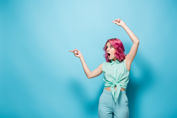 shocked young woman with pink hair pointing aside on blue background - Фото, изображение