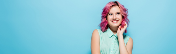 dreamy young woman with pink hair smiling and looking away on blue background, panoramic shot - Foto, afbeelding