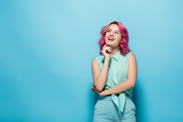 dreamy young woman with pink hair smiling and looking away on blue background - Photo, Image
