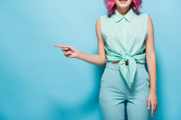vista cortada de jovem mulher com cabelo rosa apontando para o lado no fundo azul - Foto, Imagem