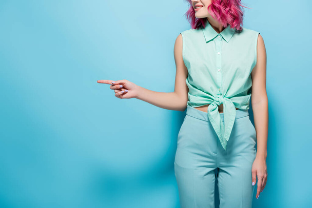vista recortada de la mujer joven con el pelo rosa señalando a un lado sobre fondo azul - Foto, Imagen