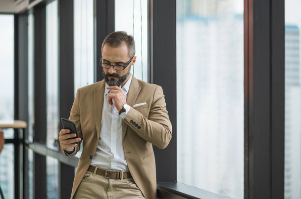 Молодий красивий бізнес бородатий чоловік стоїть пост біля вікна, використовуючи смартфон в офісному робочому місці
. - Фото, зображення