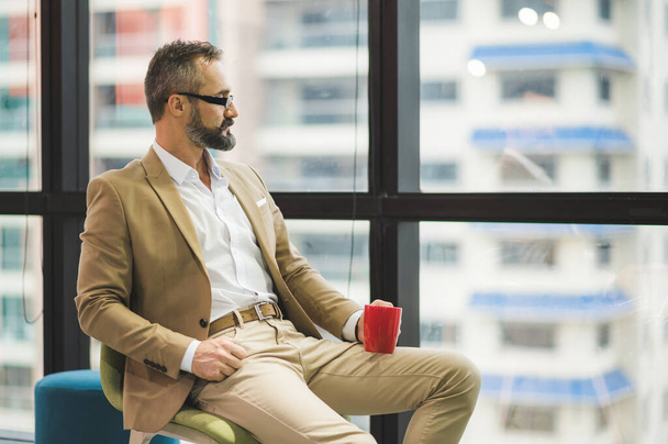 Jeune homme d'affaires beau barbu boire une tasse de café assis près de la fenêtre dans le lieu de travail de bureau. - Photo, image