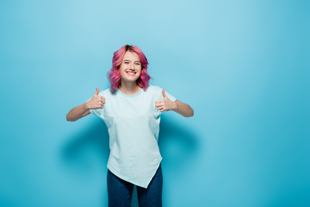 fiatal nő rózsaszín haj mutatja hüvelykujját fel kék háttér - Fotó, kép