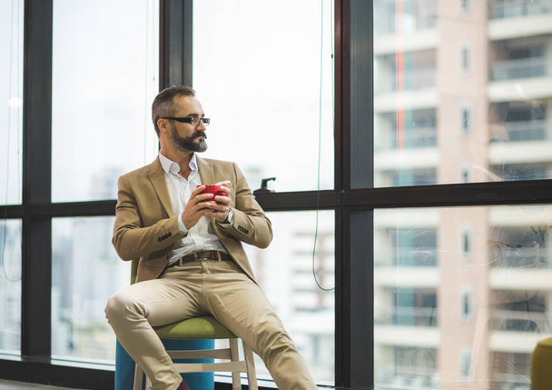 Молодий красивий бізнес бородатий чоловік п'є чашку кави, сидячи біля вікна в офісному робочому місці
. - Фото, зображення