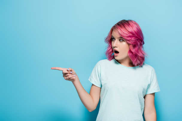 surprised young woman with pink hair pointing aside on blue background - Фото, изображение