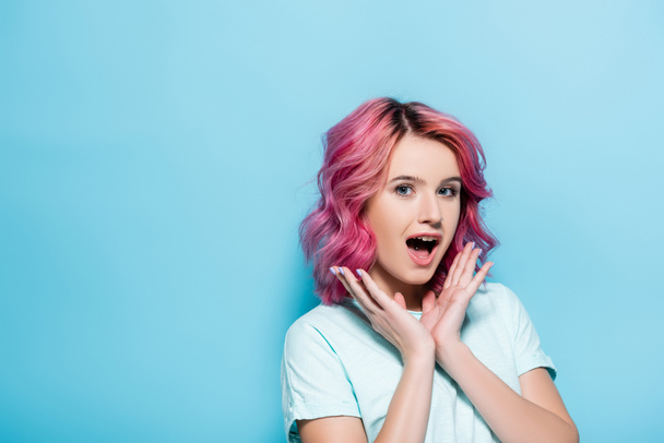 mujer joven sorprendida con el pelo rosa y las manos cerca de la cara sobre fondo azul - Foto, Imagen