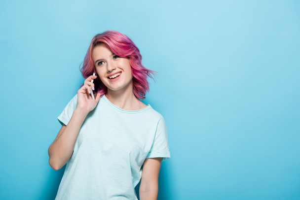 young woman with pink hair talking on smartphone and smiling on blue background - Photo, Image
